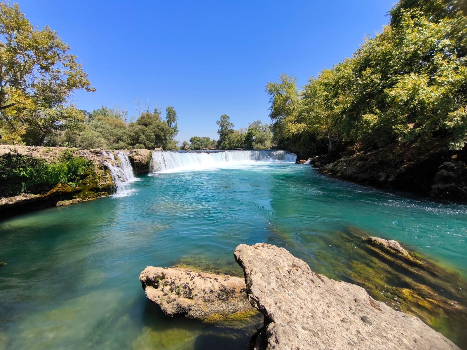 Clear turquoise river with waterfall and rocks in bright sunlight surrounded by green trees